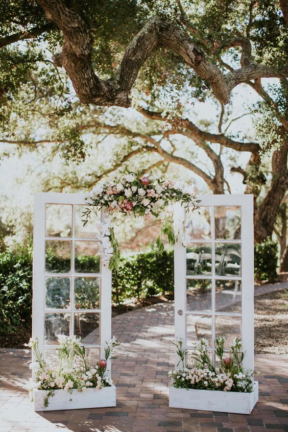 French double doors with greenery, white and pink blooms, matching blooms in planters are a great and chic wedding backdrop