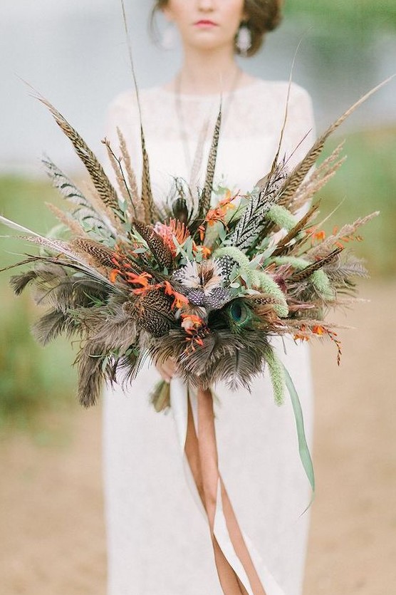 a boho fall wedding bouquet made of feathers, bright blooms, greenery and with ribbons looks unusual and catchy