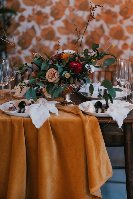 a bold fall wedding tablescape with a mustard velvet tablecloth, bold blooms and greenery, neutral porcelain and napkins