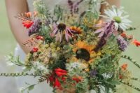 a bright wildflower wedding bouquet with greenery of various kinds, sunflowers, lilac and red touches and chamomiles is wow