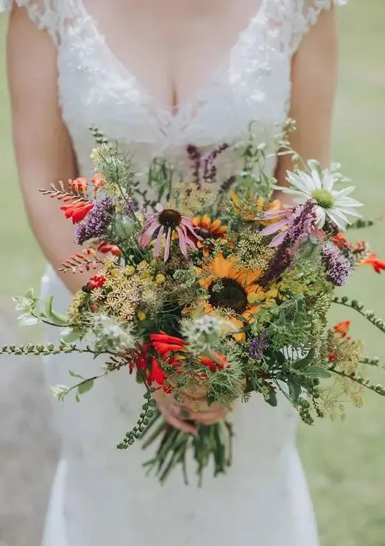 a bright wildflower wedding bouquet with greenery of various kinds, sunflowers, lilac and red touches and chamomiles is wow