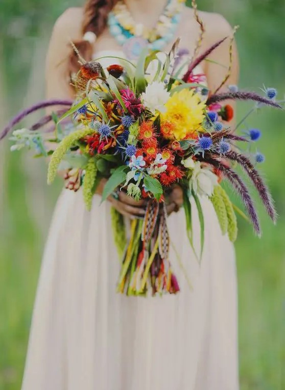 a colorful boho ethnic wedidng bouquet with orange, yellow blooms, blue thistles, lisianthus and woven ribbons