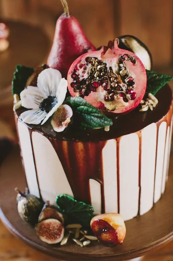a delicious fall wedding cake topped with pears, a pomegranate, figs, foliage, a white bloom and with chocolate drip