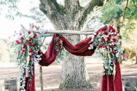 a fall wedding arch with burgundy velvet fabric, lush greenery and red roses is a beautiful idea for a refined wedding