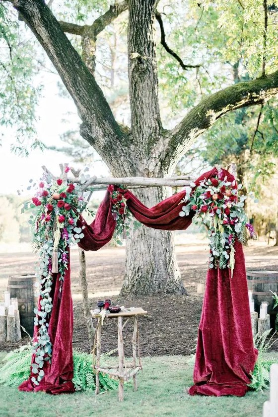 a fall wedding arch with burgundy velvet fabric, lush greenery and red roses is a beautiful idea for a refined wedding