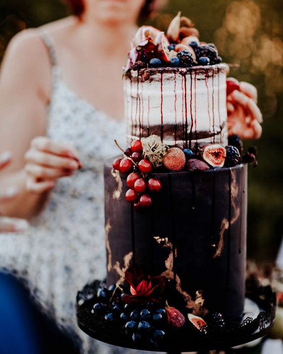 a naked and purple wedding cake with chocolate drip, gold touches, fresh fruit and berries is adorable for a fall boho wedding