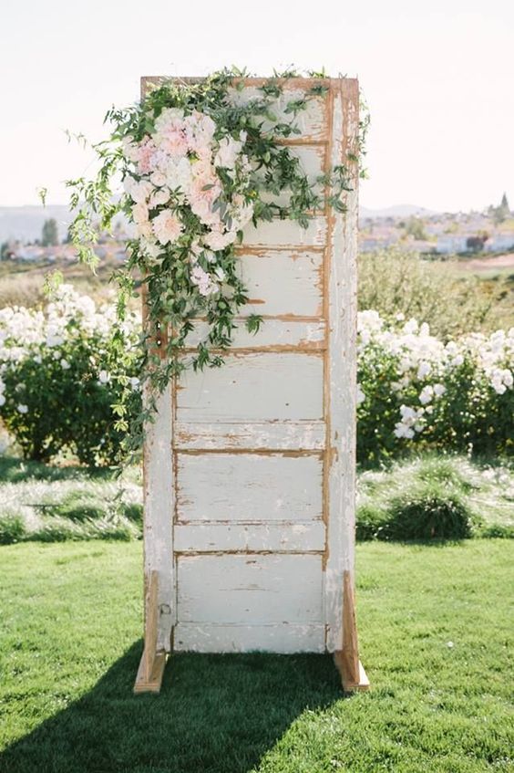 a neutral shabby chic door decorated with greenery and blush blooms on top is a lovely wedding backdrop for spring or summer
