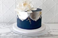 a one-tier wedding cake in navy and silver foil, with patterns and an oversized white sugar bloom on top