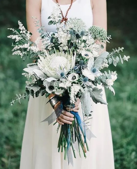 a pale and delicate wedding bouquet with blue thistles, a king protea, white anemones and herbs and eucalyptus