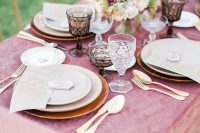 a sophisticated wedding table setting with a mauve velvet tablecloth, wooden chargers and neutral porcelain, gold cutlery and refined mauve and blush blooms