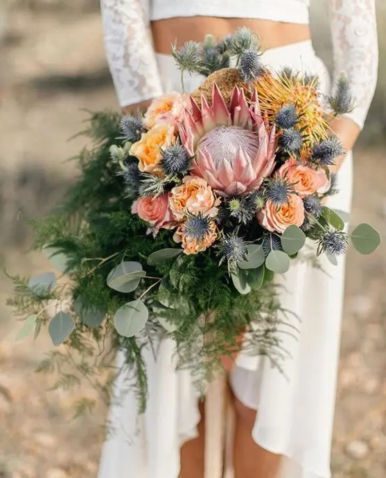 a textural cascading wedding bouquet with a king protea, peachy and yellow blooms, blue thistles and ferns and eucalyptus