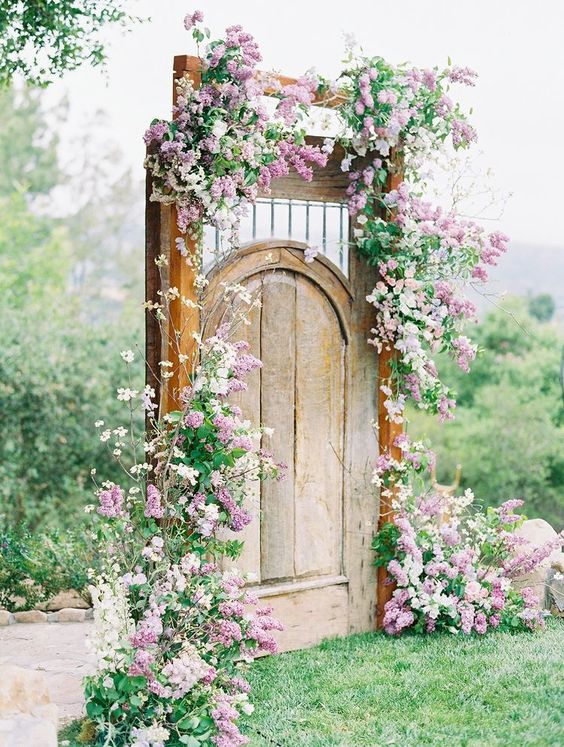 a vintage stained arched door decorated with greenery, white and lilac blooms around and on the door for a spring wedding
