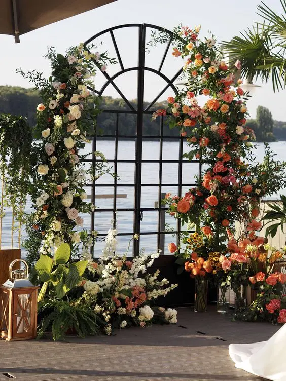 double arched French doors decorated with greenery, white and orange blooms, candle lanterns plus a water view are an amazing wedding backdrop