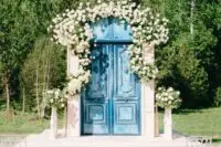refined blue shabby chic double doors decorated with neutral blooms and greenery are a lovely wedding backdrop for a chic ceremony