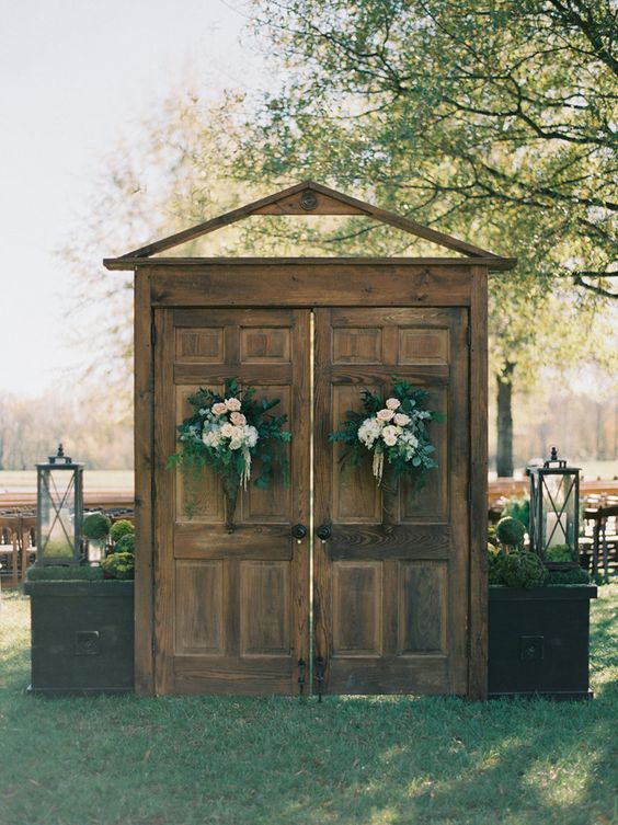 rustic stained double doors with greenery and neutral and pastel blooms plus moss topiaries and candle lanterns for a rustic wedding