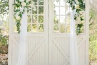 white barn doors decorated with greenery, white blooms and tulle plus pillar candles around are a chic wedding backdrop