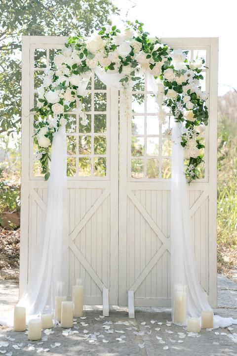 white barn doors decorated with greenery, white blooms and tulle plus pillar candles around are a chic wedding backdrop