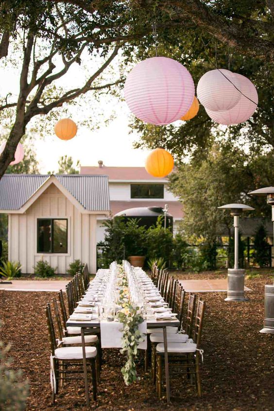 a backyard wedding reception with a neutral tablescape with a greenery and floral runner, some colorful paper lanterns over the space