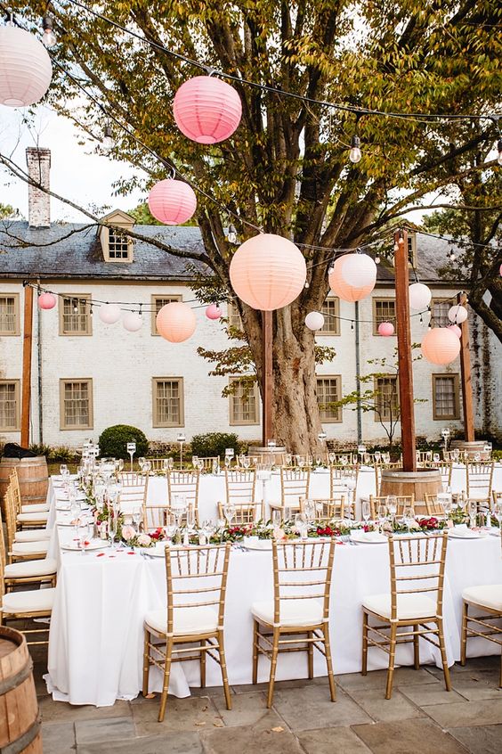 a beautiful outdoor wedding reception space with blush and red blooms and greenery, peachy, pink and neutral paper lanterns and neutral linens