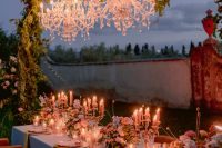 a beautiful outdoor wedding reception with a couple of crystal chandeliers hanging on greenery-covered branches over the table and lots of candles