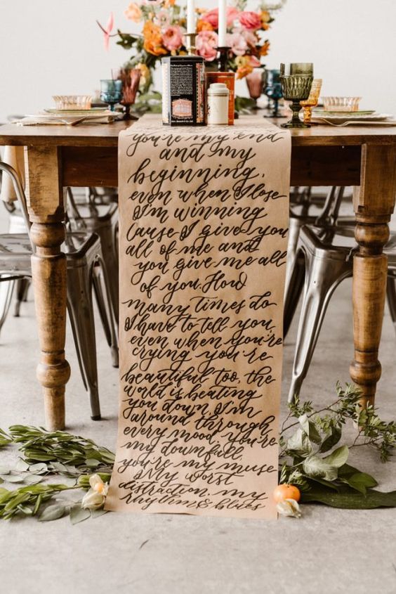 a bold wedding tablescape with a vintage table, bright blooms, a paper table runner and quotes, greenery and colorful glasses