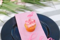 a bright tropical wedding place setting with a black plate and a pink napkin, a card and a cupckae topped with a flamingo