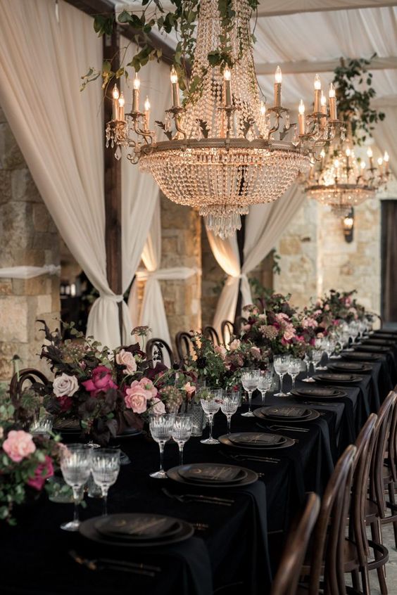 a dramatic wedding reception with a black tablescape done with blush and fuchsia blooms, dark foliage and oversized crystal chandeliers