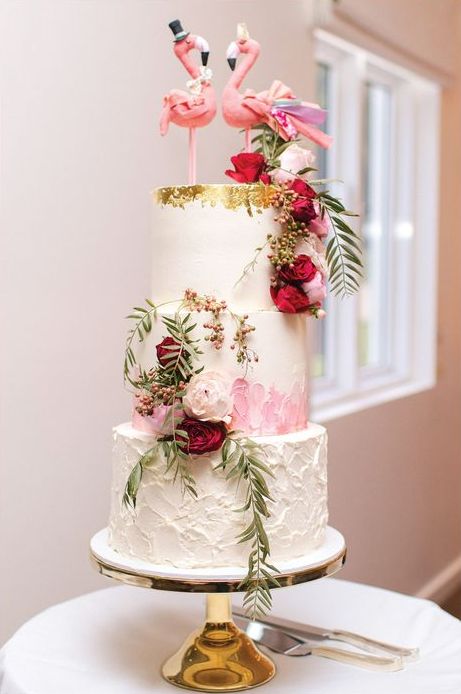 a glam wedding cake with texture and brushstrokes, with white, burgundy blooms and greeneyr and pink flamingos on top is wow