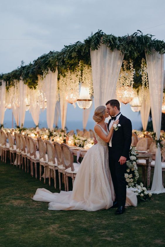 a jaw-dropping wedding reception roof covered with greenery and white blooms hanging down plus lots of crystal chandeliers over the space