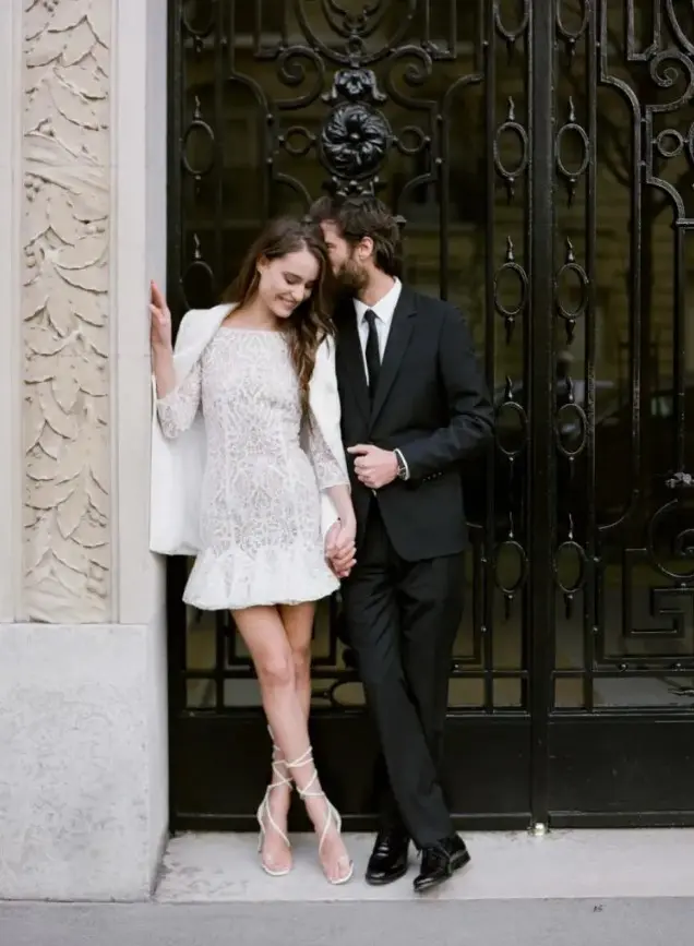 a lace high neckline wedding dress with long sleeves and a ruffled skirt is covered with a white jacket and modern lace up shoes