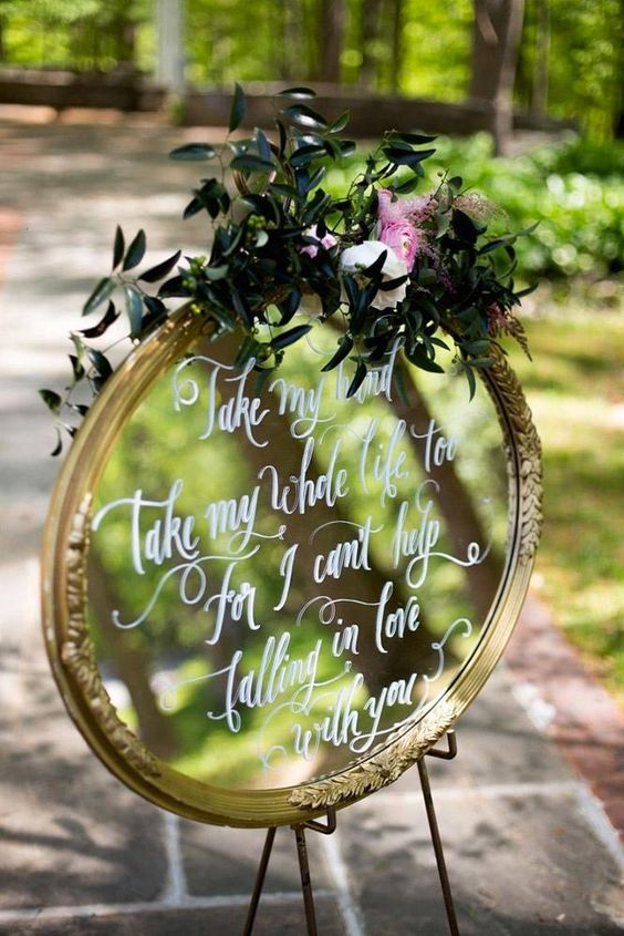 a mirror in a gold frame, with greenery and blooms plus a white calligraphy quote is a lovely decoration for a wedding