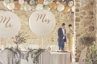 a neutral rustic wedding reception space with white textiles, balloons for accenting the couple’s chairs and arrangements of pastel paper lanterns over the space