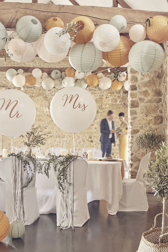 a neutral rustic wedding reception space with white textiles, balloons for accenting the couple's chairs and arrangements of pastel paper lanterns over the space