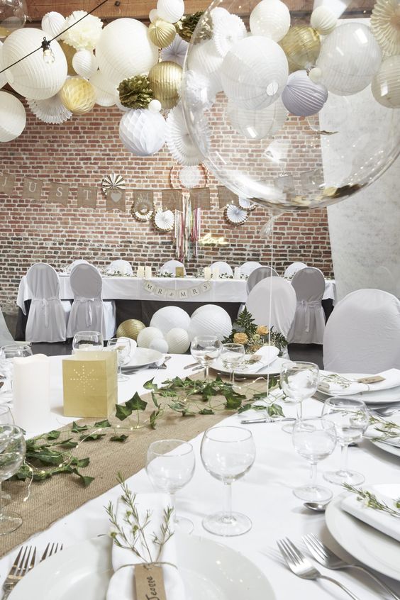 a neutral wedding reception space with white tablecloths and chair covers, with burlap table runners, greenery, string lights and white paper lanterns