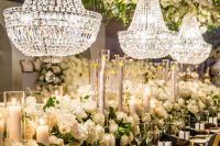 a refined and lush wedding reception with super lush neutral blooms, pillar candles and lush white florals plus crystal chandeliers over the table for a wow look