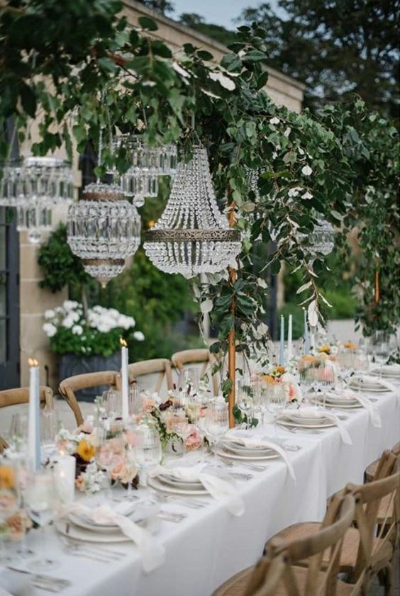 a relaxed outdoor garden wedding reception with a greenery arch over the table holding several crystal chandeliers is all glam