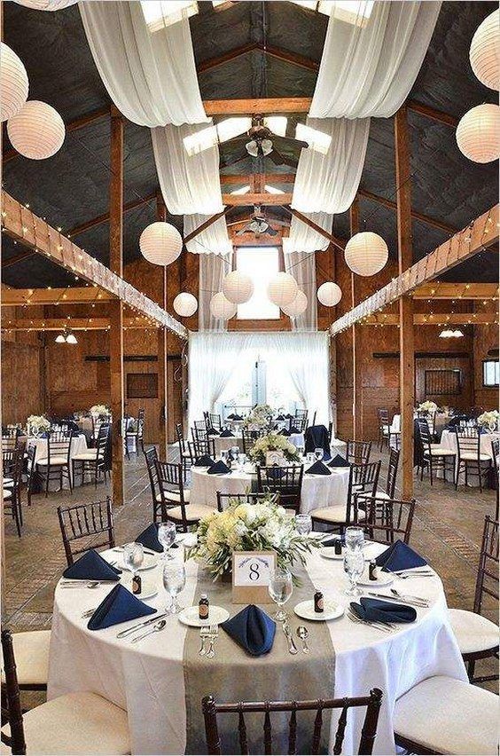 a rustic barn wedding reception decorated with neutral fabric, white paper lanterns, neutral tablescapes and navy napkins is wow