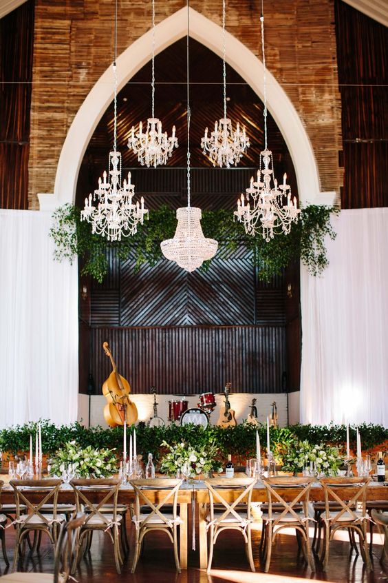 a stylish indoor wedding reception with lots of greenery on the table and over it and several pretty chandeliers to add more light to the space