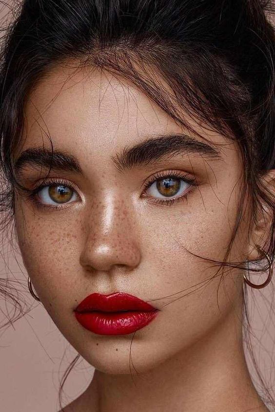 a super bold makeup with a glossy red lip, highlighted skin, trendy eyebrows and brown eyeshadows, a touch of blush on the cheeks
