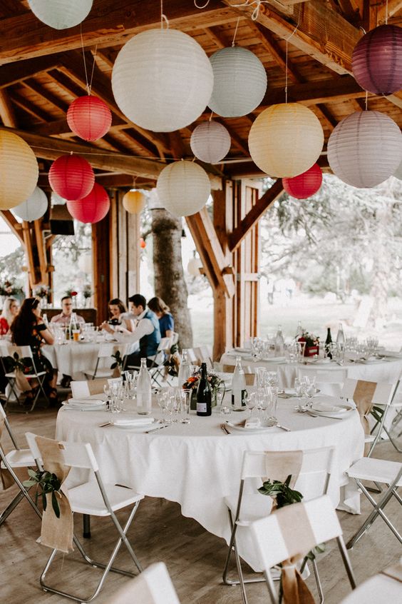 a welcoming indoor-outdoor wedding reception space with white linens, greenery and burlap chair runners, colorful and neutral paper lanterns