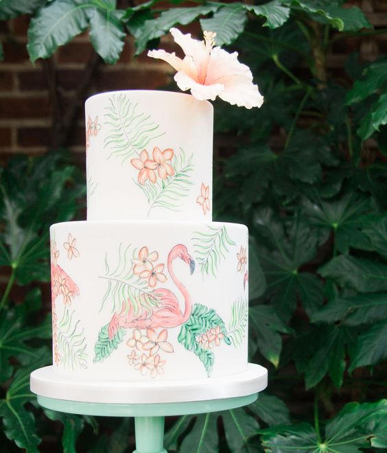a white wedding cake paitned with greenery, blooms and flamingoes and with a tropical bloom on top is a gorgeous idea