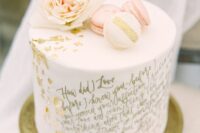 a white wedding cake with gold calligraphy quotes, with gold leaf, a blush bloom and white and pink macarons is wow