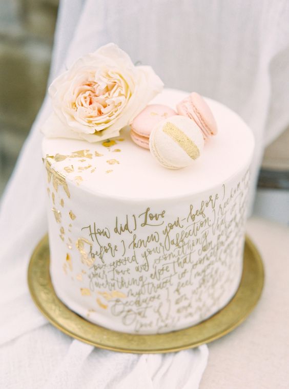 a white wedding cake with gold calligraphy quotes, with gold leaf, a blush bloom and white and pink macarons is wow