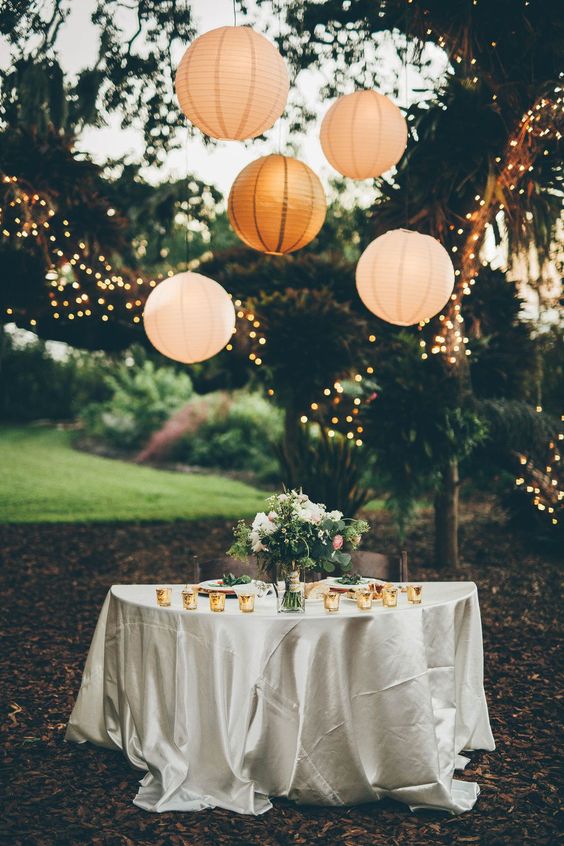 an outdoor sweetheart table with a greenery and floral arrangement, neutral paper lanterns is a lovely nook in a garden