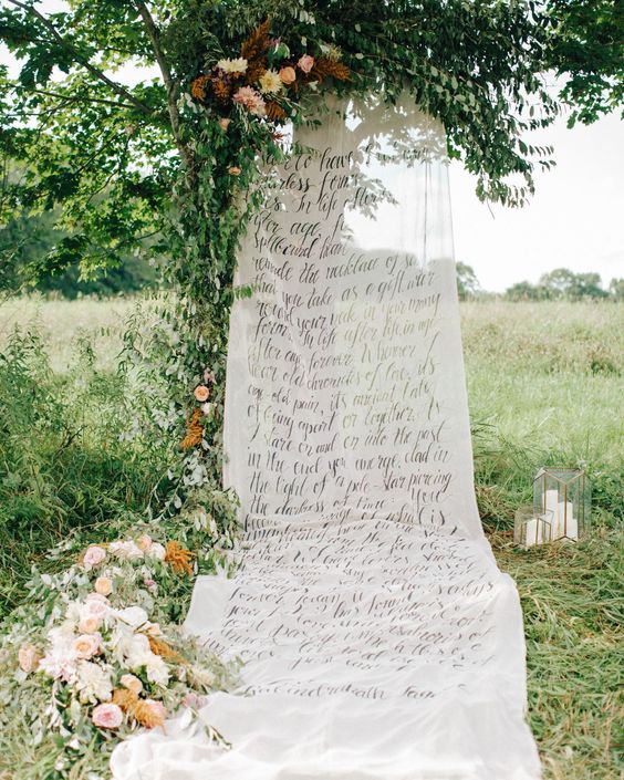 an outdoor wedding backdrop with fabric with quotes hanging on the tree, with greenery and pink and neutral blooms
