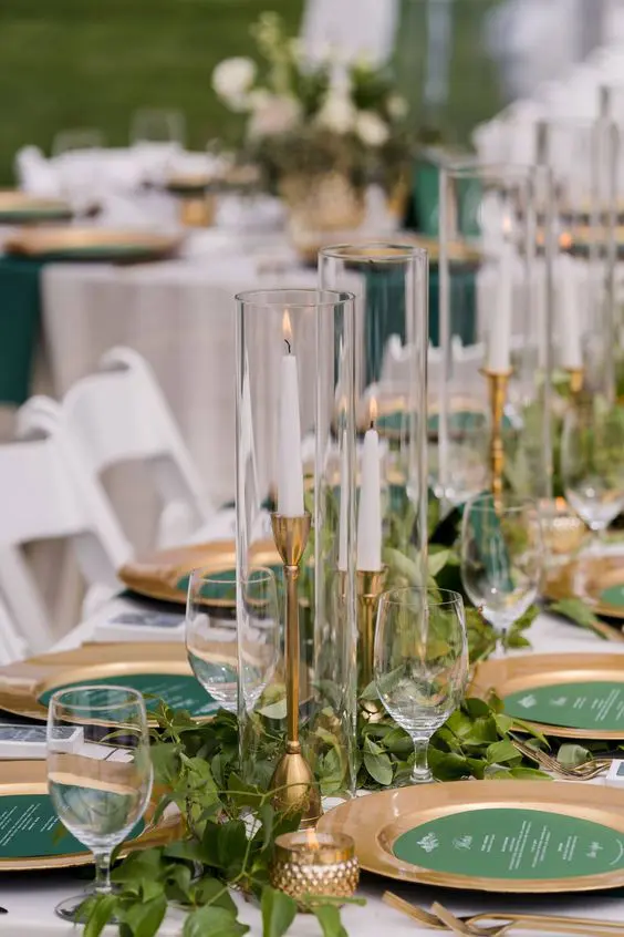 a beautiful wedding tablescape with gold placemats, candleholders and cutlery, greenery on the table and green menus is chic