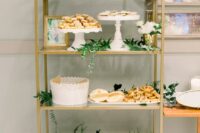 a brass shelving unit decorated with greenery and the couple’s photos is a lovely alternative to a usual wedding dessert table or an addition to it