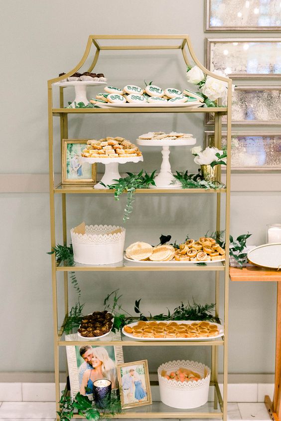 a brass shelving unit decorated with greenery and the couple's photos is a lovely alternative to a usual wedding dessert table or an addition to it