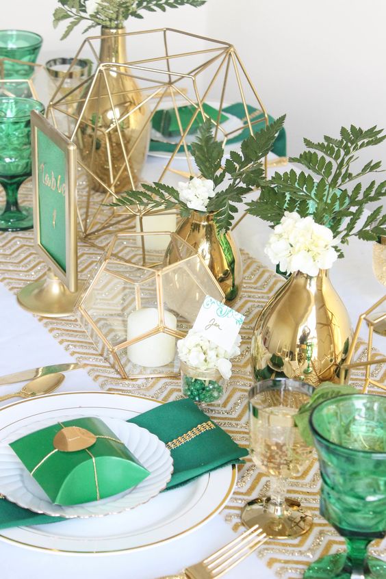 a bright and cool wedding table setting with a gold glitter runner, gold candleholders, vases and glasses, cutlery, emerald napkins and boxes