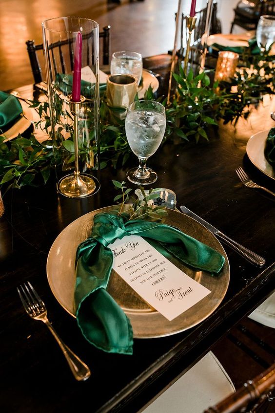 a chic emerald and gold wedding table setting with gold placemats, candleholders and cutlery, emerald napkins and greenery
